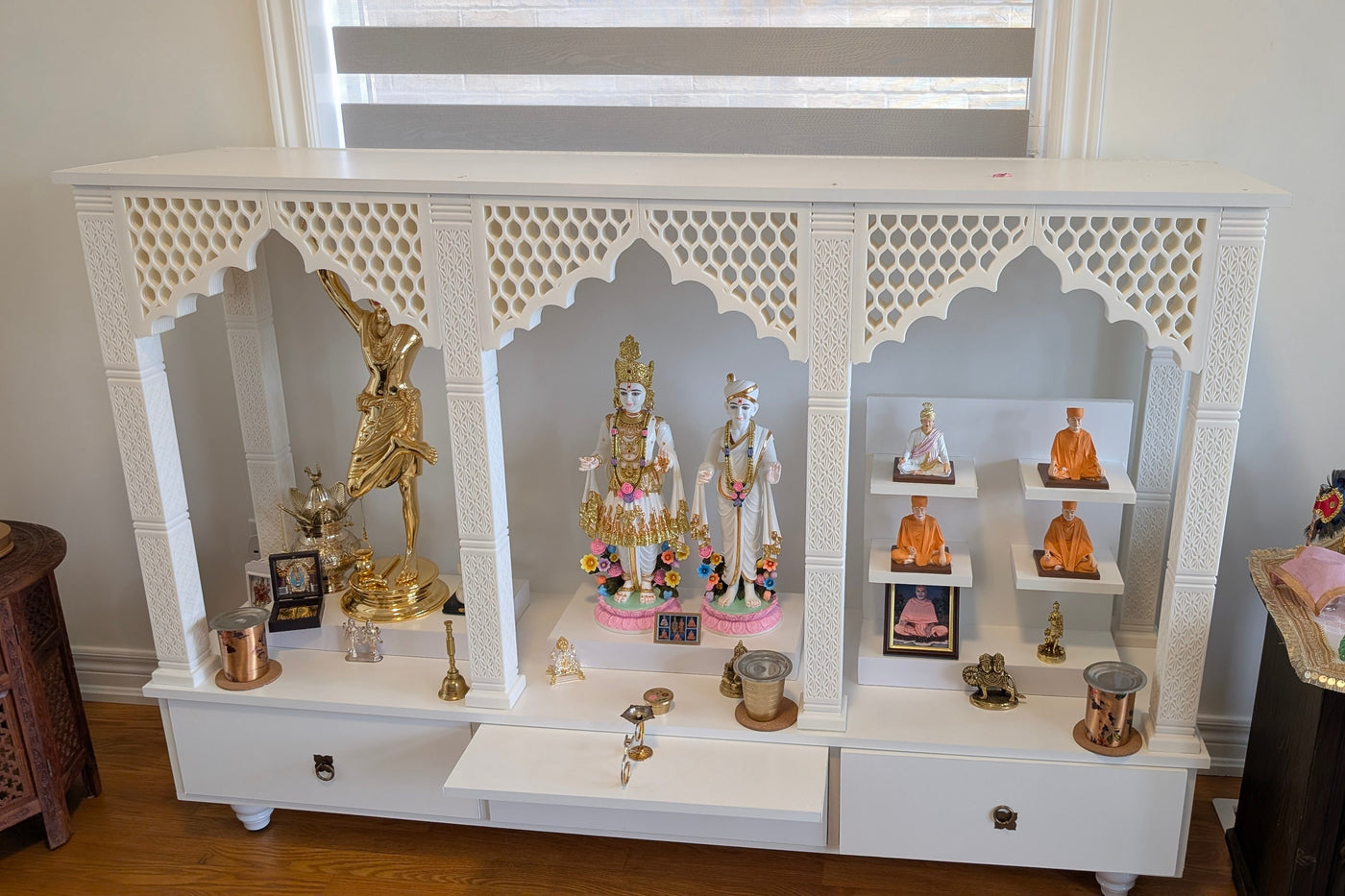 A white wooden Shiva mandir display cabinet with carved arched niches, filled with various Hindu deity statues, brass oil lamps, and decorative items, set against a light-colored wall.